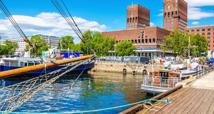 Oslos waterkant met aangemeerde zeilboten en de stenen tweelingstorens van het stadhuis onder een blauwe hemel.