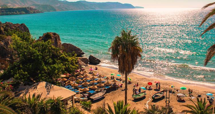 Playa con bañistas, sombrillas y barcos en el mar.