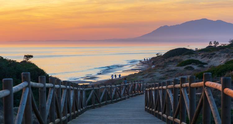 Atardecer sobre un malecón de madera y playa distante con gente.