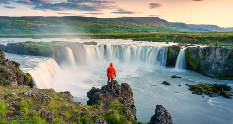 Persona de pie junto a una cascada al atardecer en medio de una exuberante vegetación.
