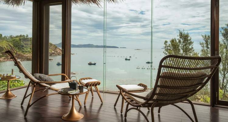 Stylish lounge chairs overlook a calm turquoise bay dotted with fishing boats through floor-to-ceiling windows.