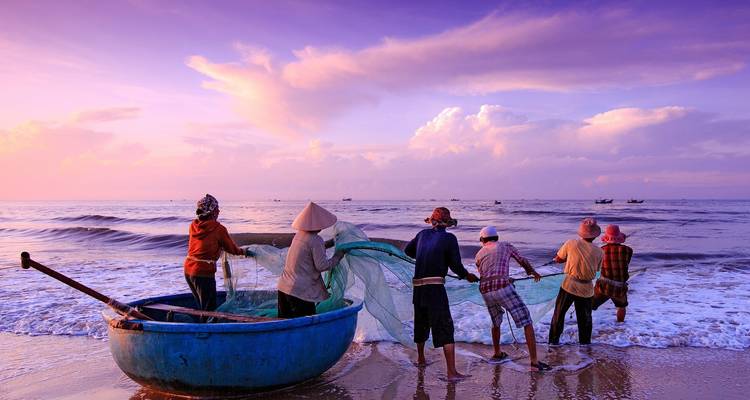 Fishermen in conical hats pull in a large net at dawn on a gentle surf beach bathed in purple light.