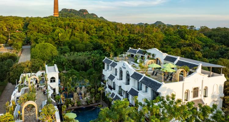Charming white villa resort with rooftop terraces sits amid dense green forest below a tall pagoda tower.