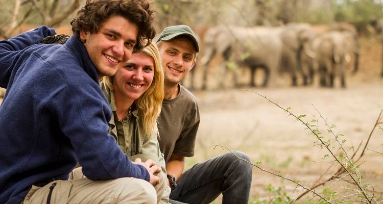 Three smiling young travellers crouch near a safari track with a herd of elephants blurred in the background.