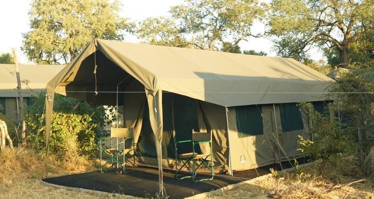 Simple canvas safari tent with two camp chairs on a small platform beneath trees.