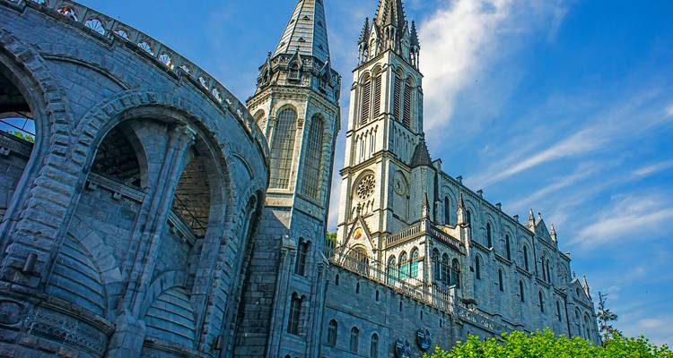 De stenen bogen en torens van de Basiliek van Lourdes steken dramatisch af tegen een levendige blauwe hemel.