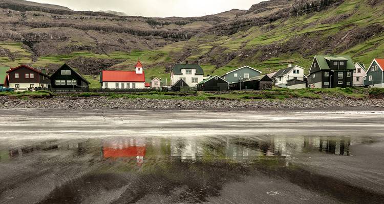 Rangée de maisons colorées et petite église reflétées dans le rivage de sable noir mouillé sous des falaises vertes abruptes.