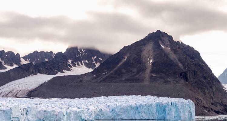 Schroffe dunkle Berge, teilweise in tiefe Wolken gehüllt, ragen über eine zerklüftete Gletscherfront und eisiges Wasser empor.