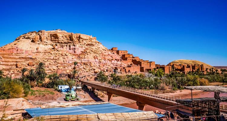 La Kasbah de Ait Benhaddou se alza en una colina sobre un río seco y palmeras de oasis bajo un cielo despejado.