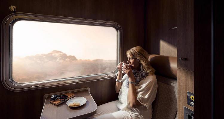Een vrouw die aan een warme drank nipt in een luxe treinkabin terwijl het landschap in de dageraad voorbij het raam glijdt.