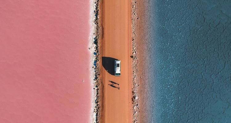 Drone view of a vehicle on a causeway dividing a pink salt lake and a blue lagoon with long shadows.