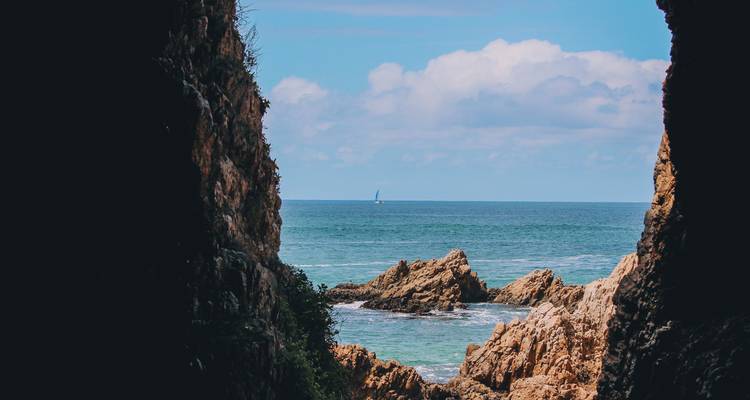 Un paisaje marino enmarcado a través de una grieta en la costa rocosa revela un velero en el horizonte.