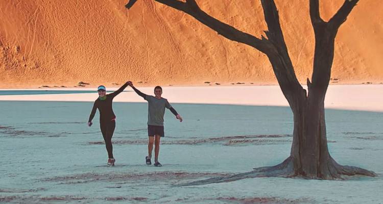 Couple holding hands walks beneath a lone tree on the white pan of Namibia's Dead Vlei with towering orange dunes behind.