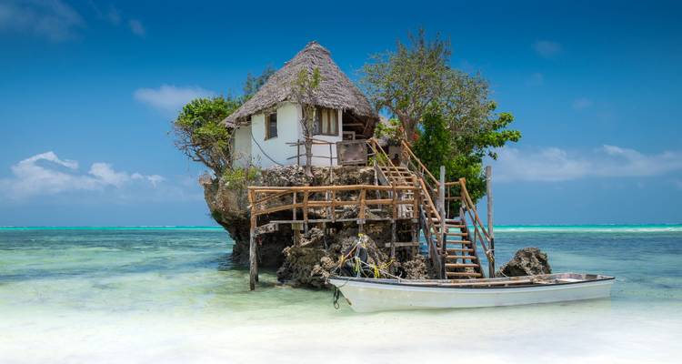Restaurante costero único en una roca alta con un barco cerca.