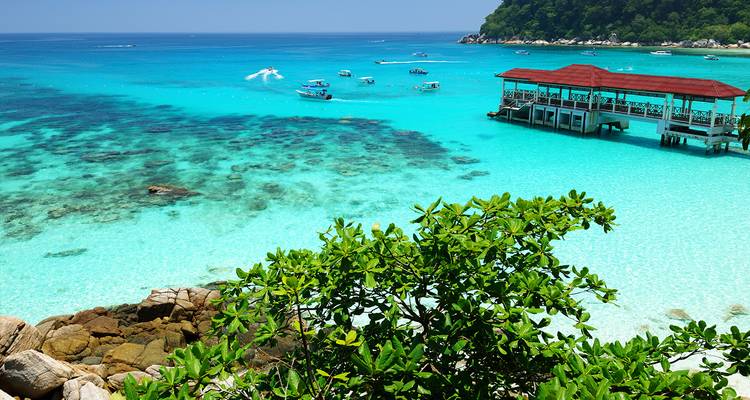 Bahía tropical turquesa con arrecifes de coral, pequeñas embarcaciones y un muelle de madera bajo un cielo azul despejado.