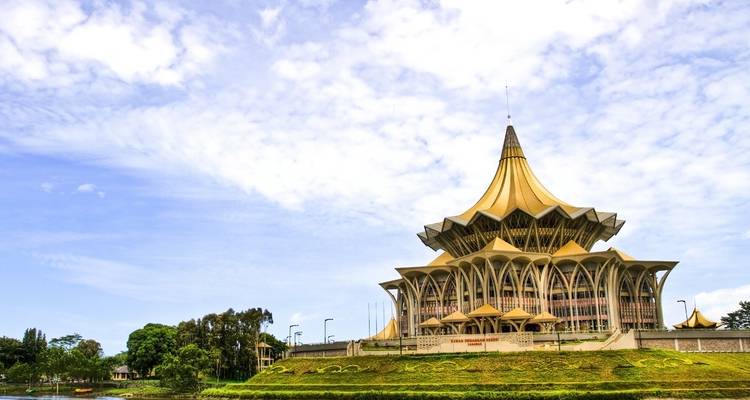 Bâtiment distinctif de l'Assemblée législative de l'État de Sarawak au toit doré situé au bord d'une rivière sous un ciel bleu.