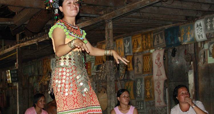 Danseur autochtone de Sarawak vêtu d'une tenue traditionnelle se produisant à l'intérieur d'une maison commune en bois tandis que des spectateurs regardent.
