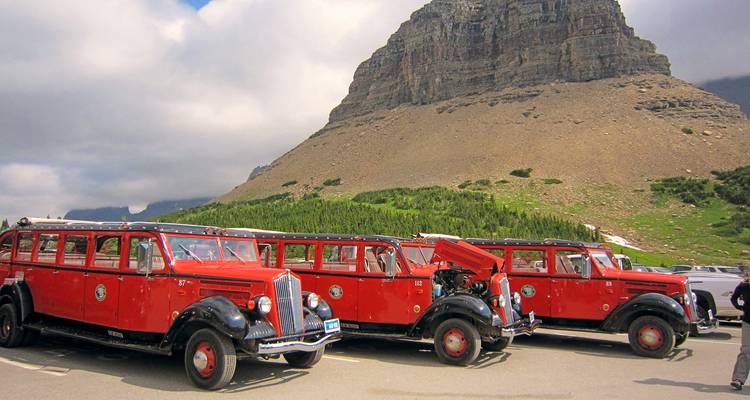 Fila de autobuses clásicos rojos de tour vintage estacionados debajo de una ladera rocosa de montaña.