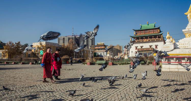 Los monjes caminan por una plaza mientras las palomas revolotean alrededor de los edificios históricos del templo.