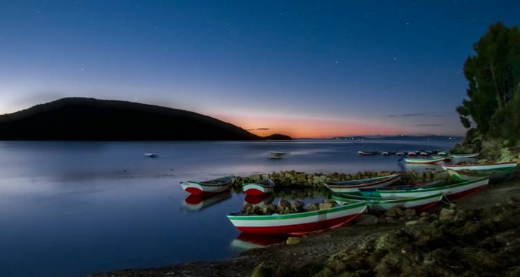Les bateaux de pêche peints reposent sur les eaux calmes du crépuscule tandis que le soleil se couche derrière les collines sombres et que les premières étoiles apparaissent dans le ciel dégagé.