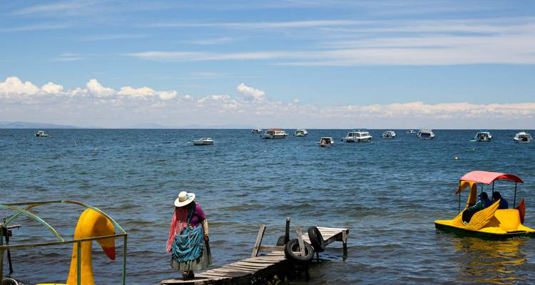 Una mujer con ropa andina tradicional camina por un pequeño muelle de madera mientras innumerables botes se mecen en el enorme lago azul.