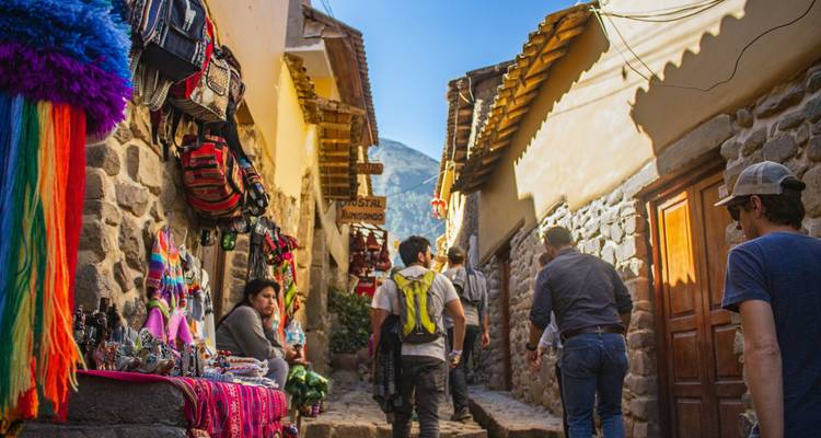 Los viajeros pasean por un estrecho callejón de piedra lleno de puestos de artesanías coloridas y textiles colgantes en el mercado de un pueblo andino.