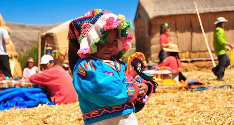 Smiling child in vibrant embroidered clothing holds a woven doll on the golden reeds of a floating island.