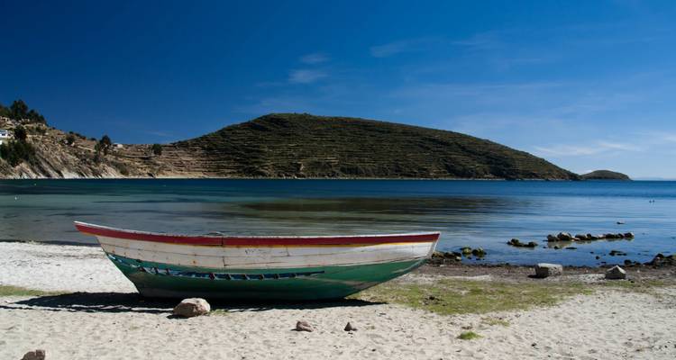 Painted wooden boat rests on a tranquil beach of Lake Titicaca with a verdant island hill in the background.
