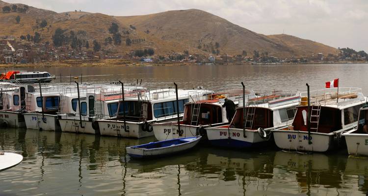 Row of excursion boats tied together at a pier on Lake Titicaca with arid hills and townscape of Puno beyond.