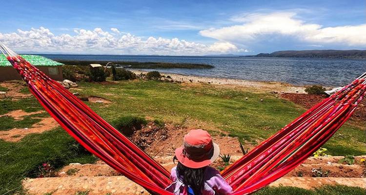 Traveller in a red hammock relaxes facing the tranquil expanse of Lake Titicaca and a distant cloud-filled horizon.