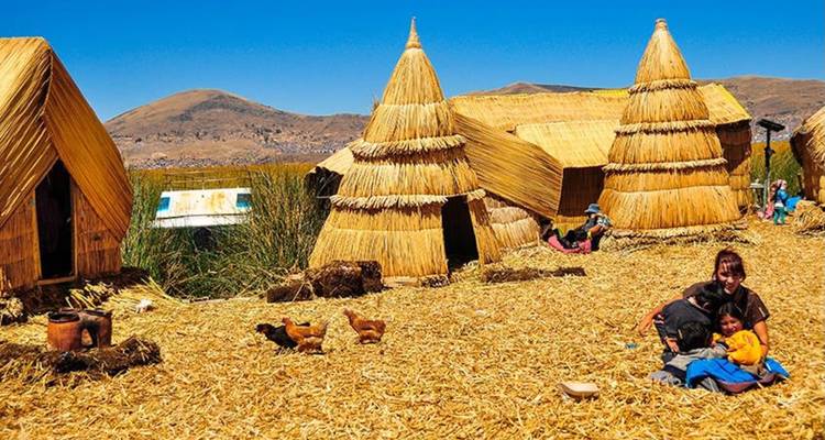 Families and chickens move about a settlement of reed huts on a floating island backed by dry Andean hills.