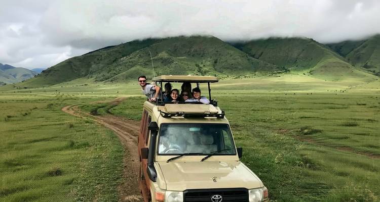 Open-roof safari vehicle with smiling travellers driving across open grassland toward green hills under low cloud.