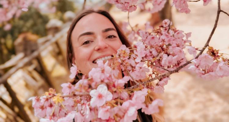 Glimlachende vrouw poseert achter delicate roze kersenbloesems in soft focus.