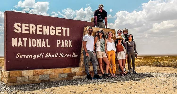 A cheerful group poses beside the Serengeti National Park entrance sign on a partly cloudy day.