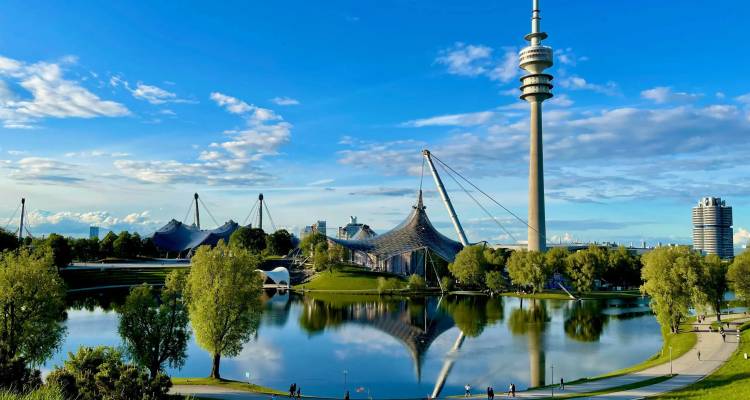 Helder zomertafereel van Munichs Olympiatore weerspiegeld in het rustige meer met blauwe lucht.
