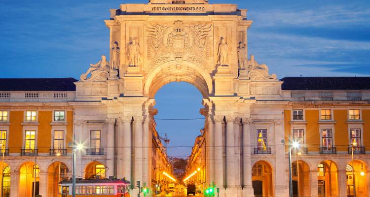 Triumphal arch with evening lights and a tram passing in front.