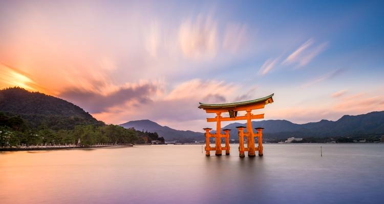 Famous torii gate at sea with mountains in the background during sunset.