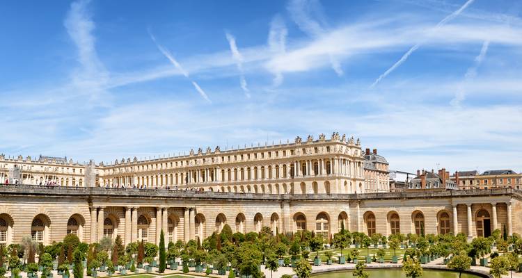 Weitläufige Ansicht der Fassade des Schlosses Versailles mit gepflegten Parterre-Gärten unter gestreiftem blauem Himmel.