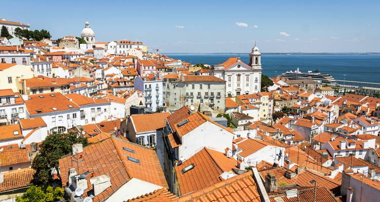 Vue panoramique sur les toits orange d'Alfama descendant vers le Tage et un navire de croisière amarré en bas.