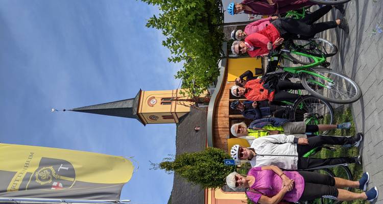 Un groupe de cyclistes seniors pose avec des vélos de location vert vif devant une petite église villageoise et des bannières.