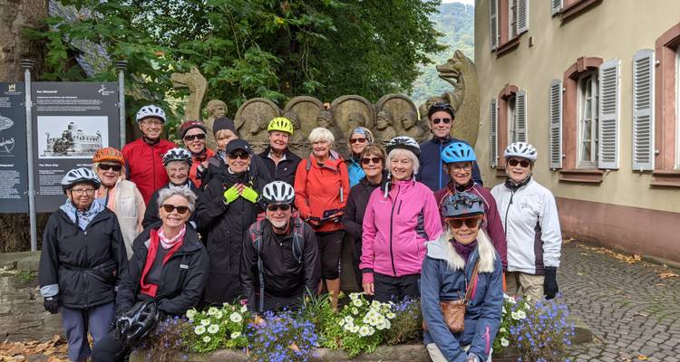 Un grand groupe de cyclistes vêtus de vestes colorées et de casques sourit pour une photo à côté d'une sculpture de dragon en pierre.