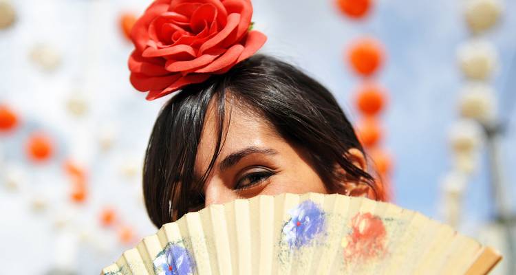 Femme portant un accessoire capillaire en forme de rose rouge se cachant de manière ludique derrière un éventail décoré.