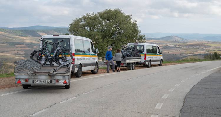 Las furgonetas de apoyo con remolques para bicicletas esperan en un costado de la carretera con vistas mientras los ciclistas charlan junto a ellas.