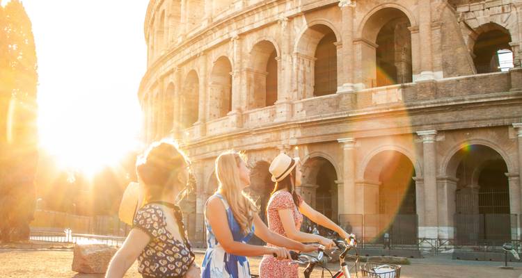 Trois jeunes femmes avec des vélos admirent le Colisée éclairé par le soleil, le flare de l'objectif créant une lueur chaleureuse.