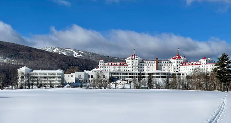 Snow-covered field stretches toward the grand Mount Washington Hotel set against forested mountains.