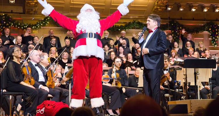 Festive orchestra concert with Santa Claus taking center stage in a decorated concert hall.