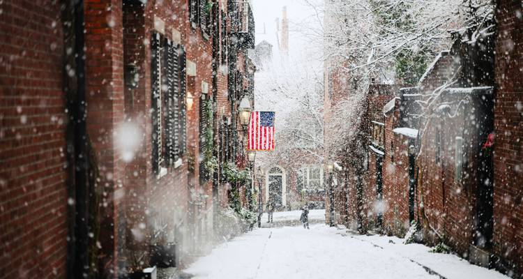Narrow brick alley in Boston blanketed with falling snow and an American flag hanging mid-street.