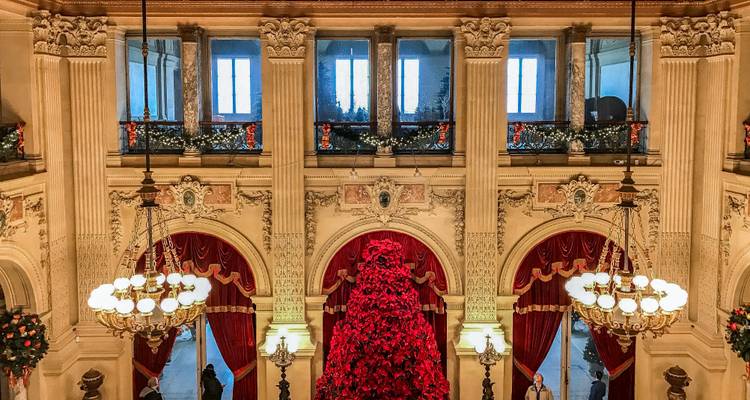 Lavishly decorated Gilded Age mansion interior with towering red poinsettia Christmas tree and chandeliers.