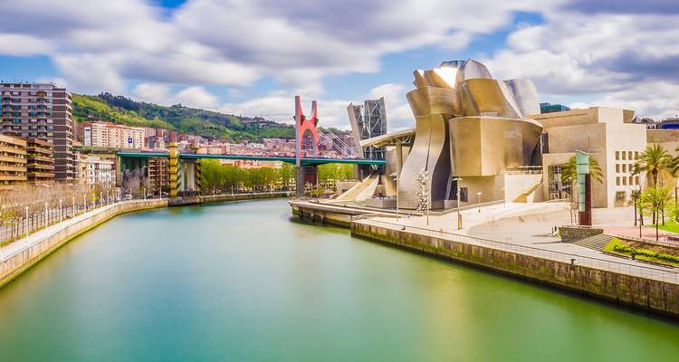 El reluciente Museo Guggenheim de Bilbao se curva junto al río Nervión bajo un cielo parcialmente nublado.