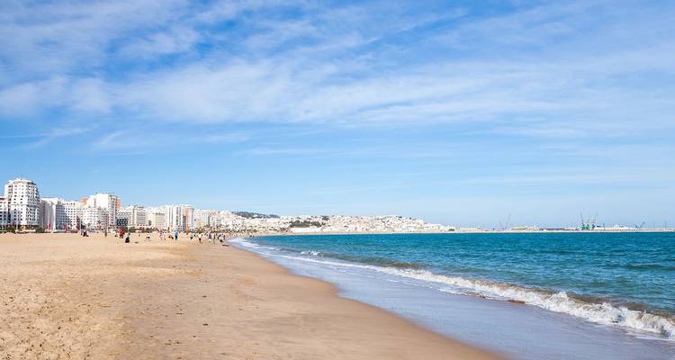 Una amplia playa urbana de arena blanca se curva junto a un mar turquesa con un horizonte de edificios blancos a lo lejos.